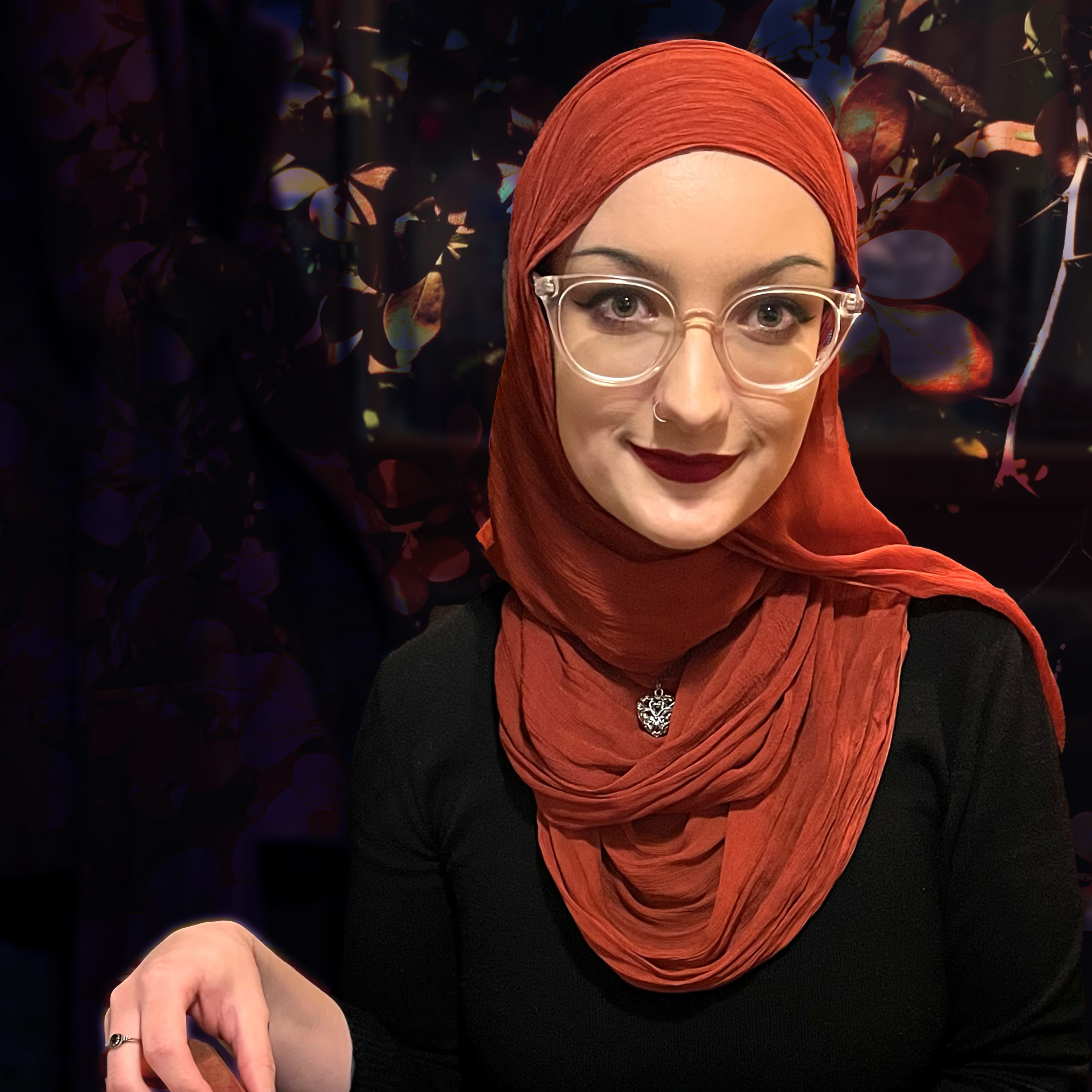 A 20-something-year-old woman wearing a dusty-red-coloured hijab looks at the camera with her head tilted. She is seated and smiling at the person taking her picture. The background is obscured. The image is black-and-white until it's hovered over. She has big green-ish eyes, and one nostril pierced. She is wearing dramatic makeup with a red lip, sharp eyeliner and thin brows.